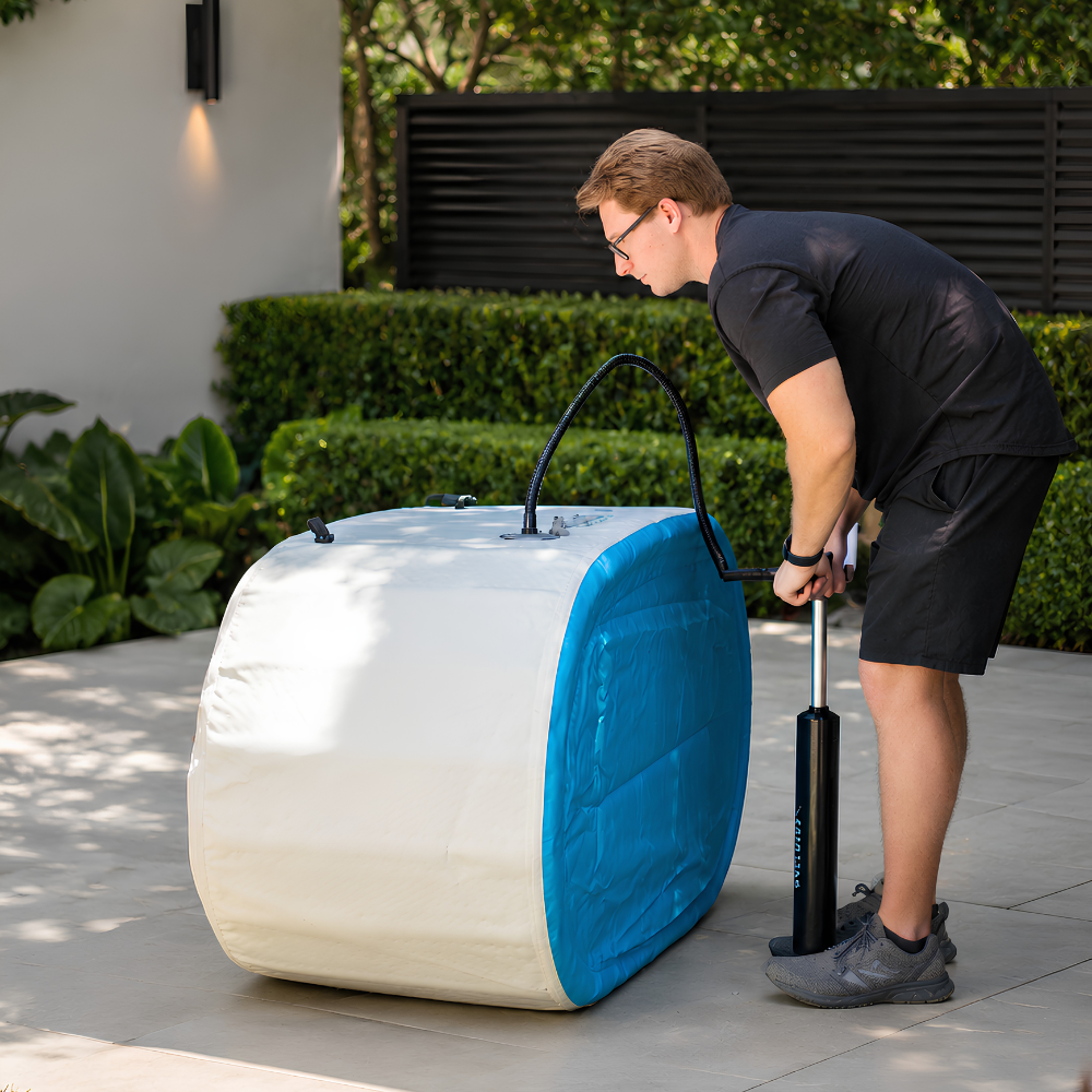 A man in a black t-shirt and shorts is using a manual air pump to inflate a RevX Inflatable Cold Plunge Tub Deluxe