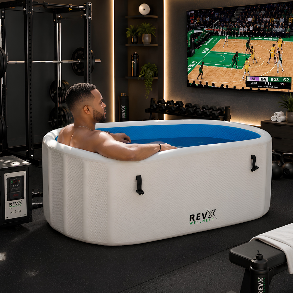 A man relaxes in a white RevX Inflatable Cold Plunge Tub Deluxe, set in a modern gym environment with a large TV showing a basketball game in the background.