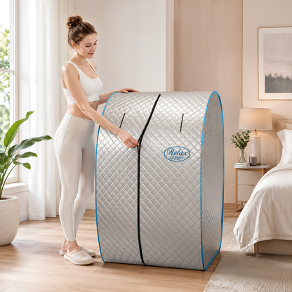 A woman in a white sports bra and leggings stands next to a portable Relax Silver Elite Far Infrared Sauna with reflective silver exterior, portable seated design, patented infrared heating technology, and compact home wellness setup in a bright bedroom setting.