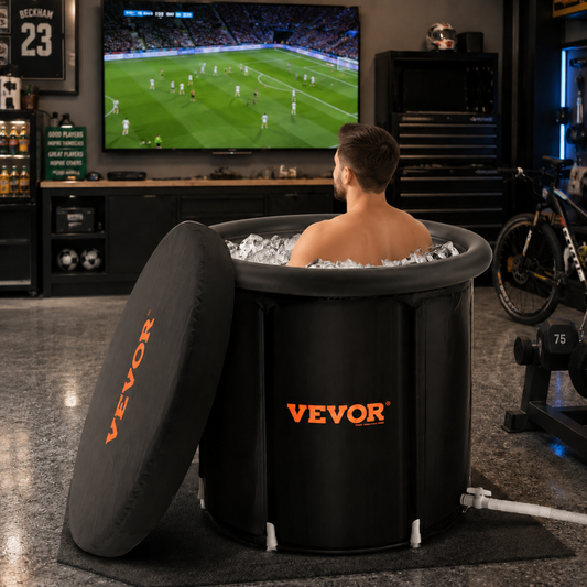 A man sits in a black VEVOR inflatable hot tub filled with ice, watching a soccer game on a TV in a dark home gym or garage setting.